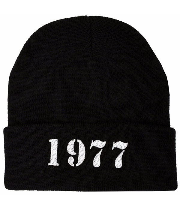 Black knitted punk beanie hat with wide folded cuff featuring bold white 1977 lettering embroidered in a distressed stencil-style font.